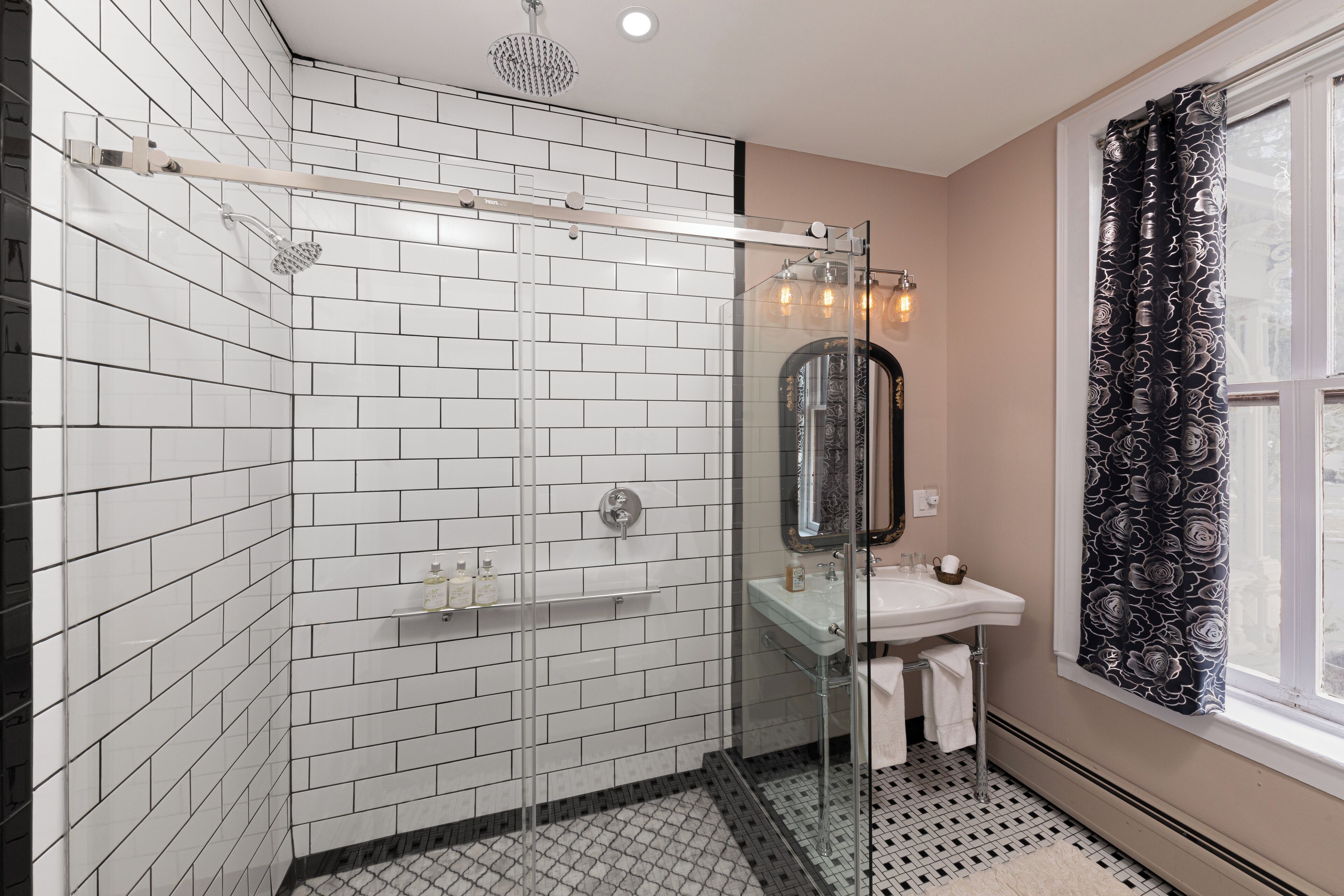 Ein modernes Badezimmer mit weißen, rechteckigen Fliesen, einer Glasdusche, einem Glaswaschbecken und einem Fenster mit gemusterten Vorhängen.