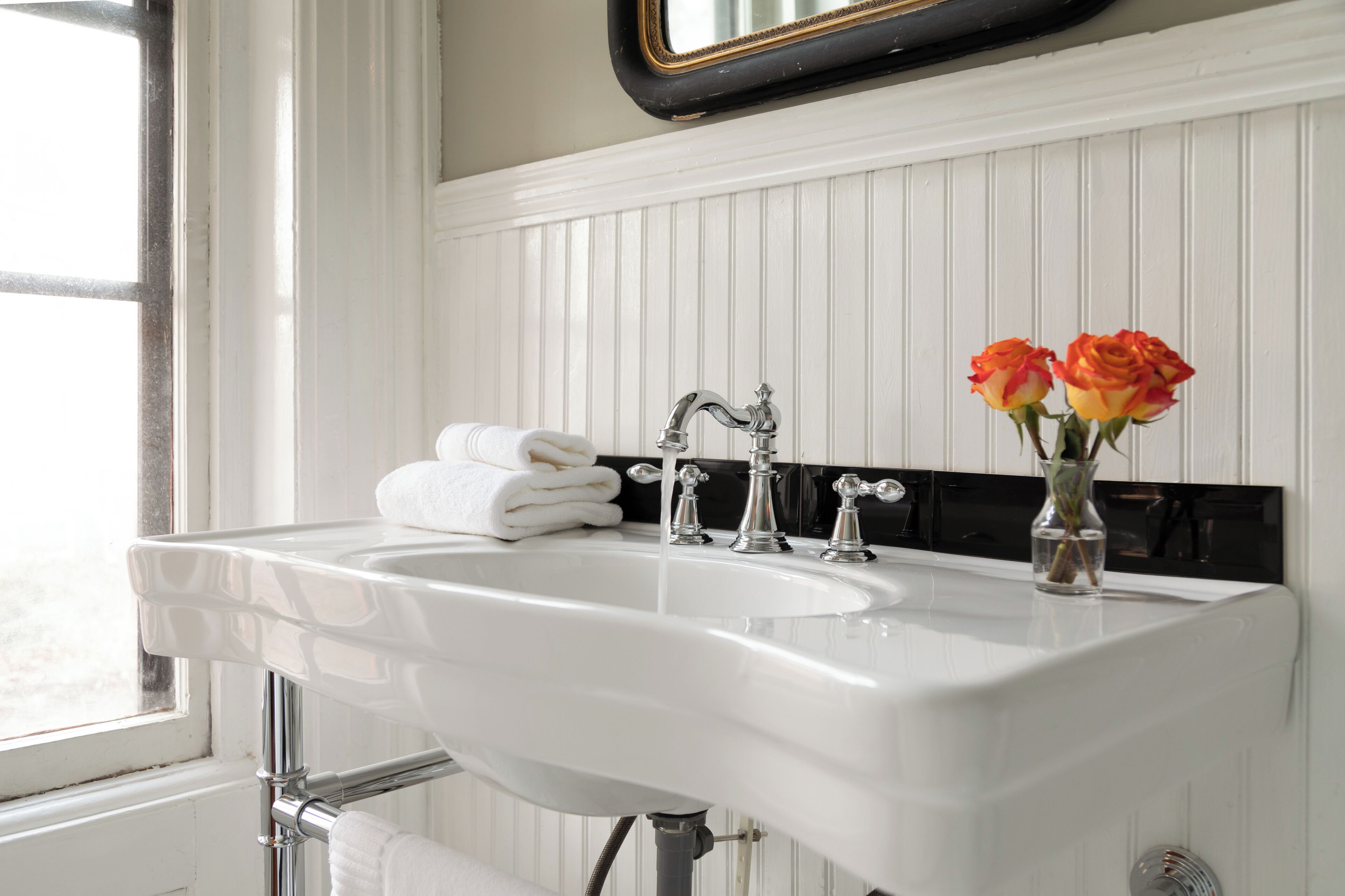 A white bathroom sink with two faucets, a mirror above it, and a vase with roses on the countertop.