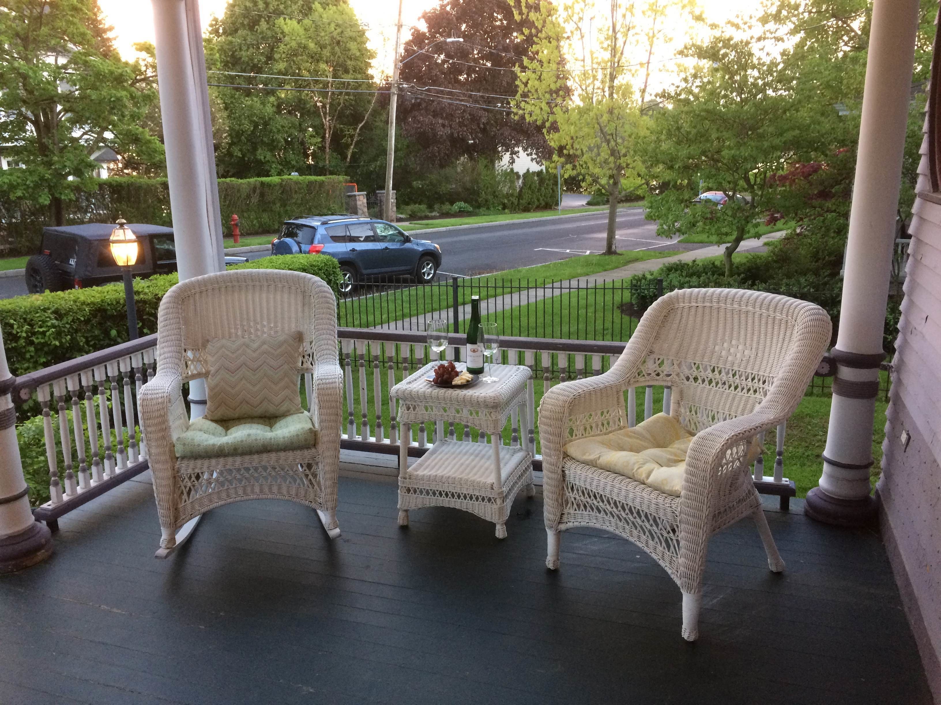 A porch with two wicker chairs, a small table with a bottle and glass, and a view of a street with cars and greenery.