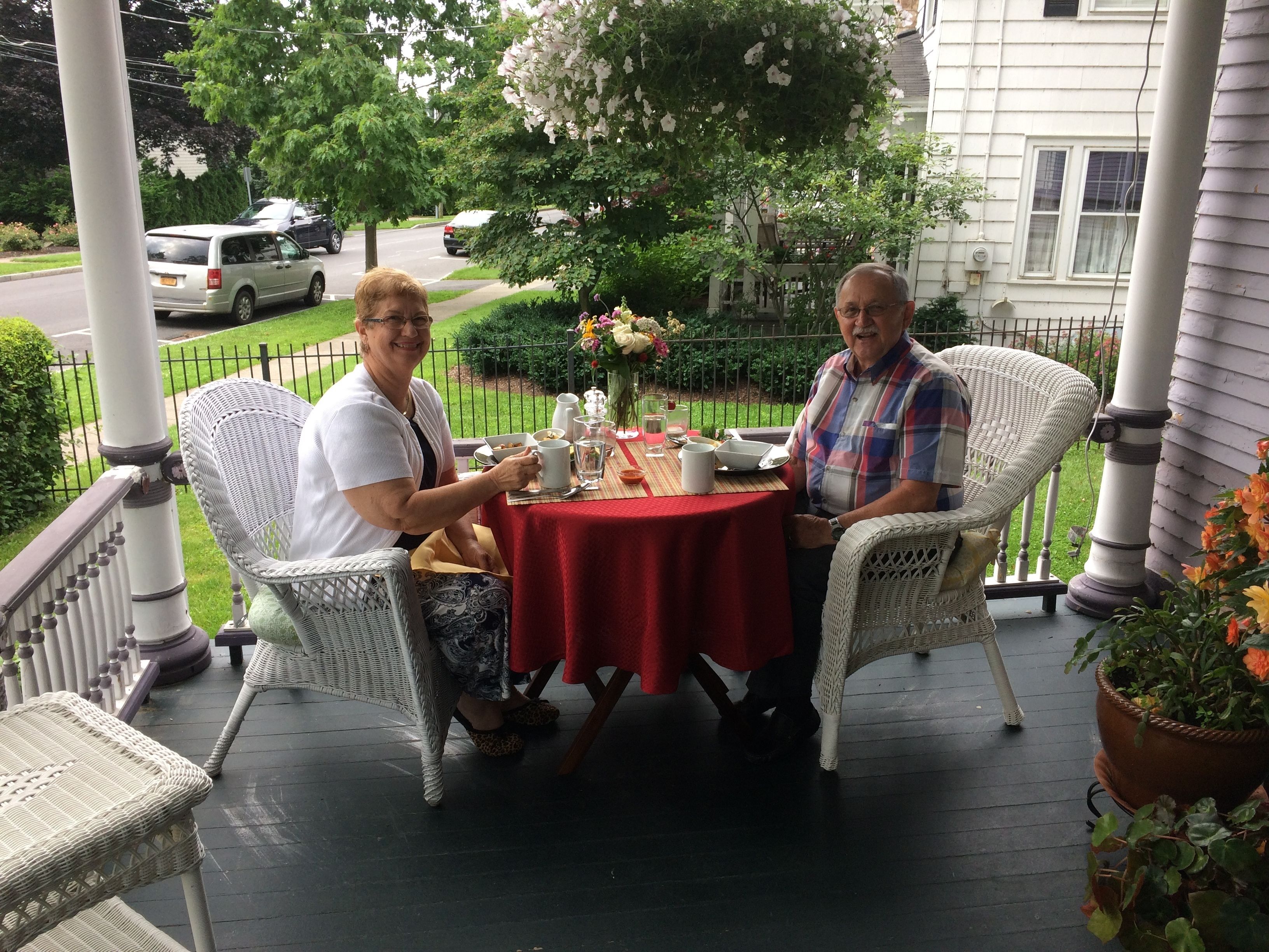 Two people seated at a round table with a red tablecloth on a porch.