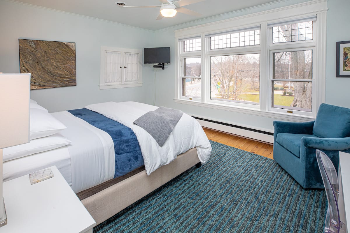 A bedroom with a bed, a blue chair, a TV mounted on the wall, and a window with a view of the outside.