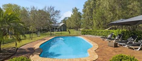 A rectangular swimming pool with a brick border, surrounded by a brick patio. There are lounge chairs and a covered area with an umbrella.