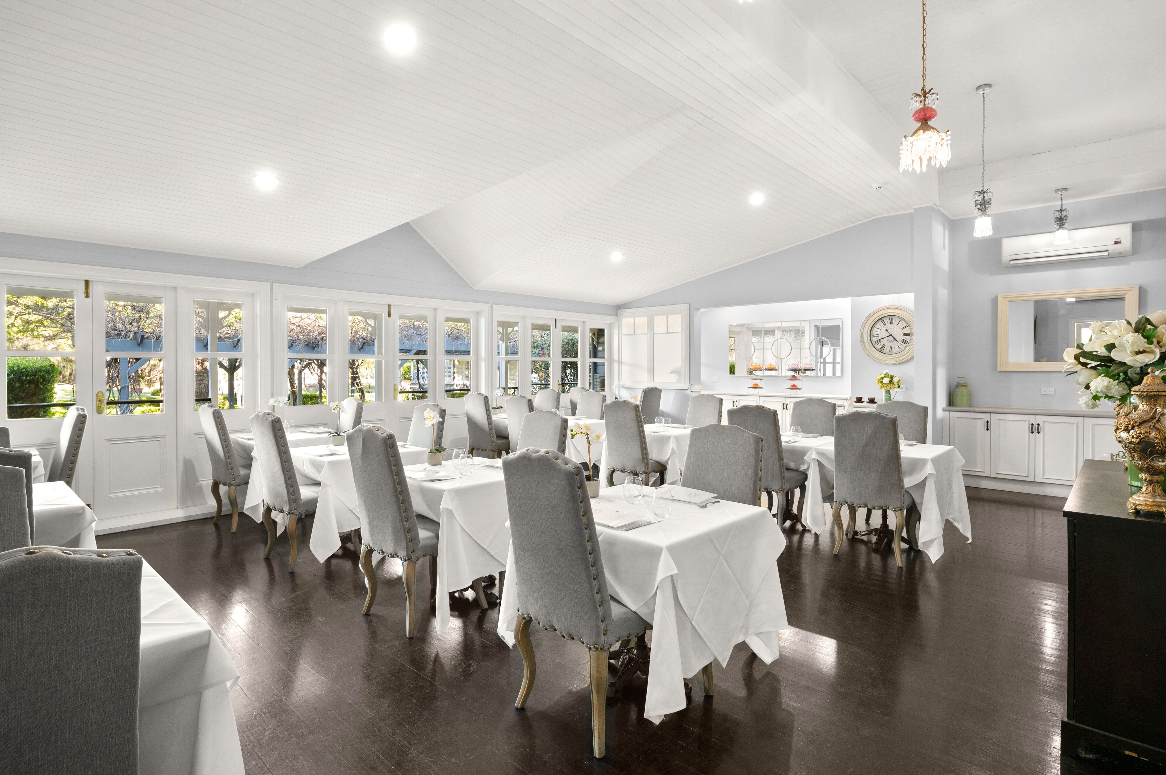 Un vaste espace dédié à la salle à manger, avec des tables et des chaises blanches, de grandes fenêtres et, au fond, une cuisine moderne.
