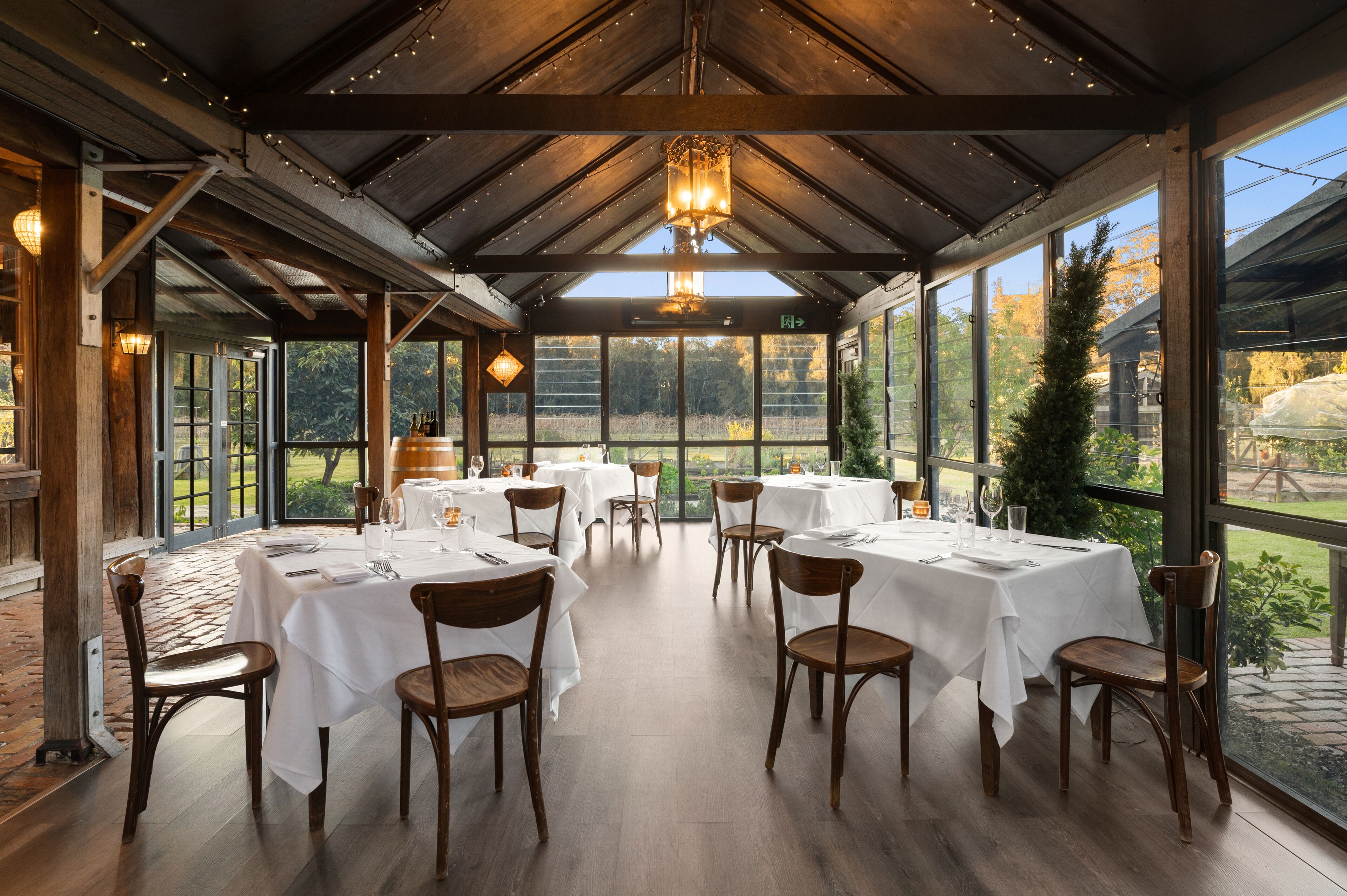 A restaurant with multiple tables set for dining, large windows offering a view of the outdoors, and a wooden ceiling structure.