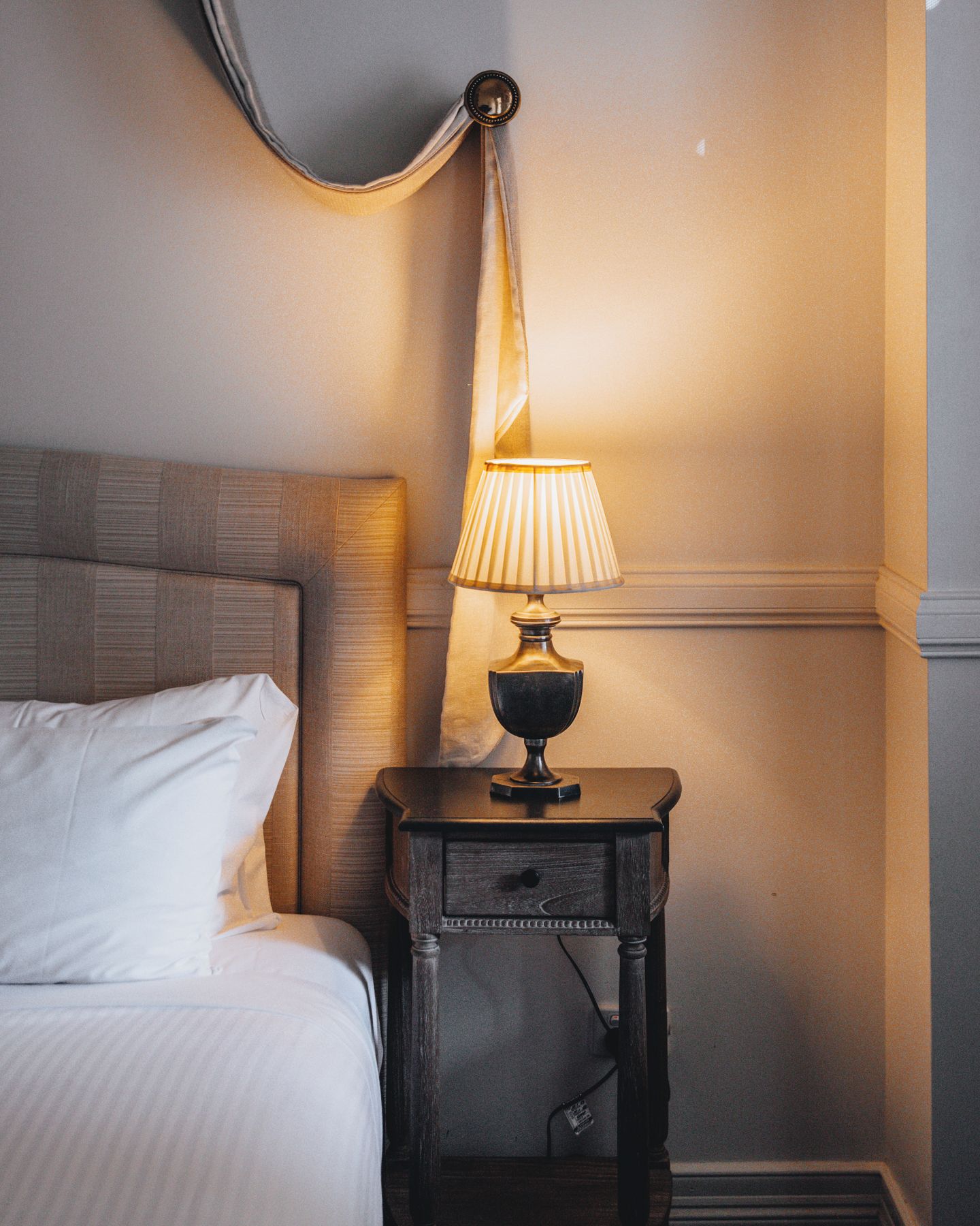 A neatly made bed with white linens, a dark wooden nightstand with a lamp, and a wall-mounted light fixture.