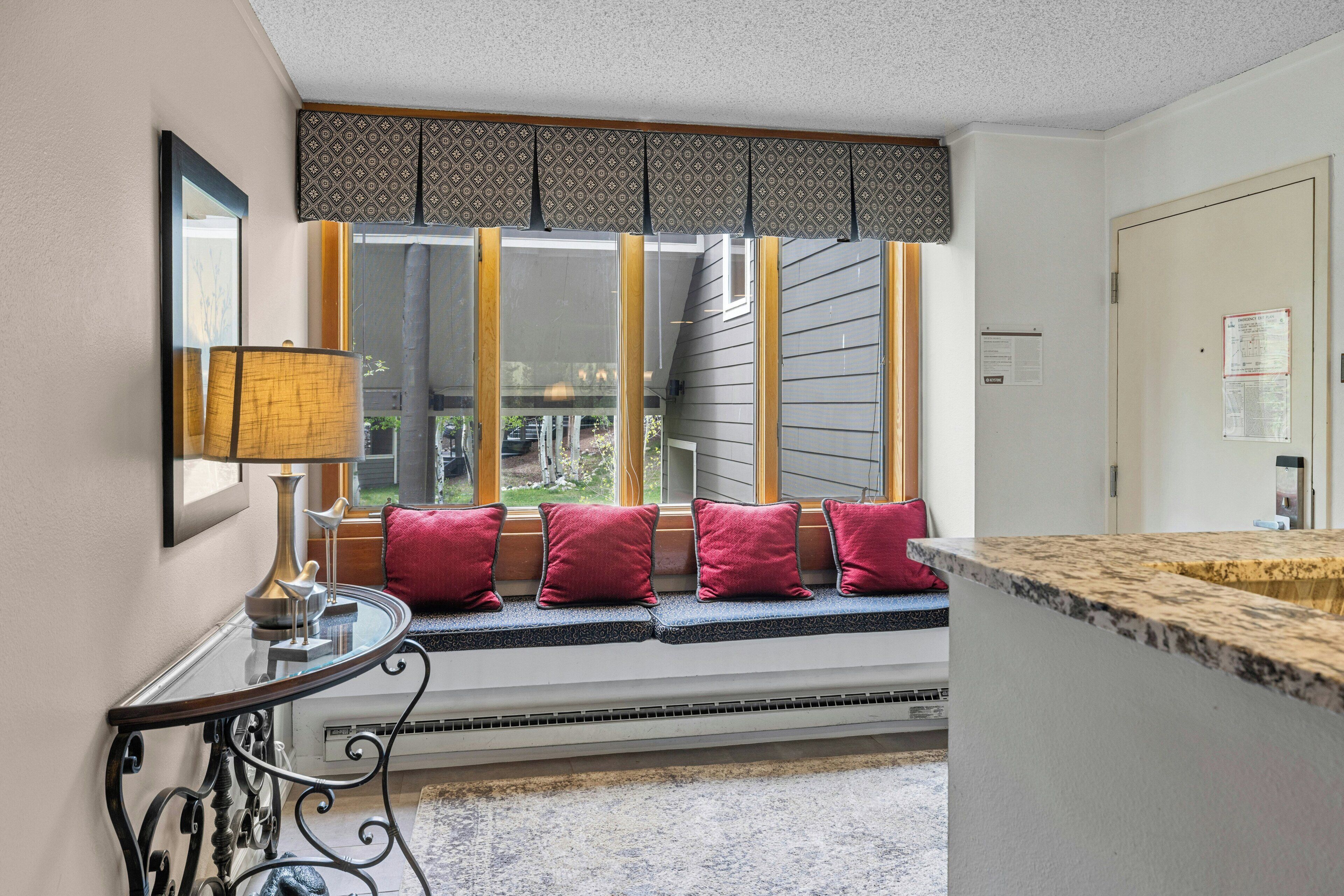 A room with a window seat, a table with a lamp, and a door with a bulletin board.