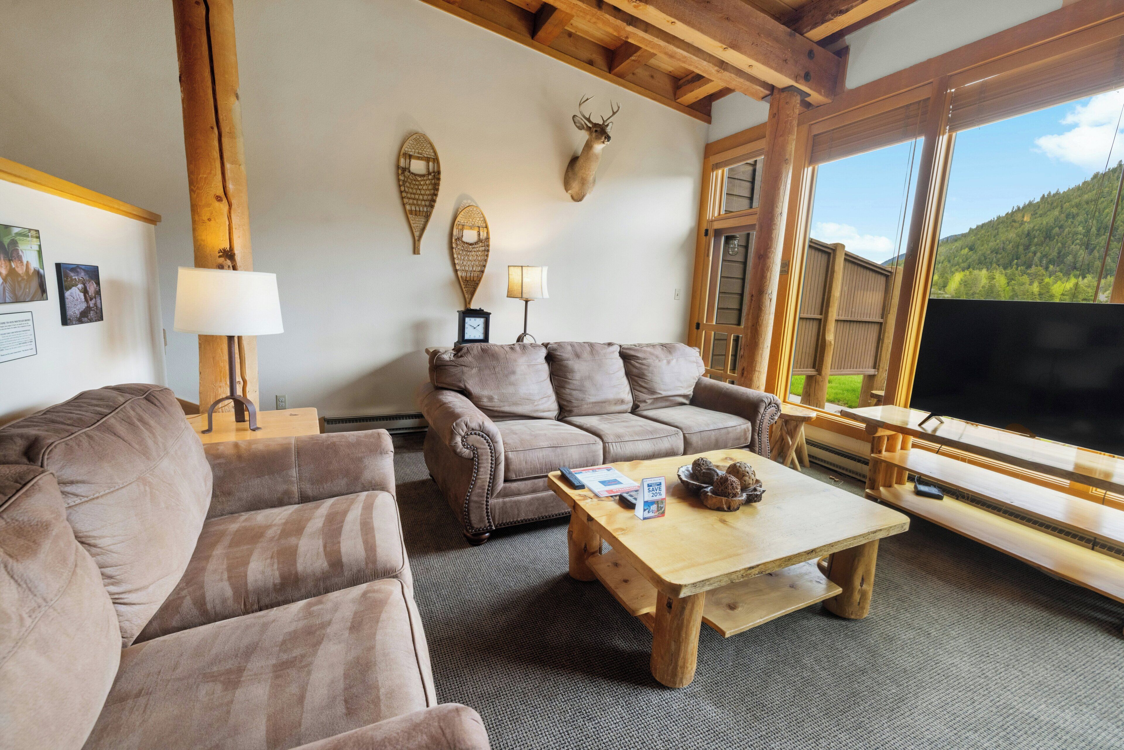 A cozy living room with a brown sofa, a wooden coffee table, and a mounted deer head. There are two wall-mounted lamps, a framed picture, and a large window with a view of a mountain.