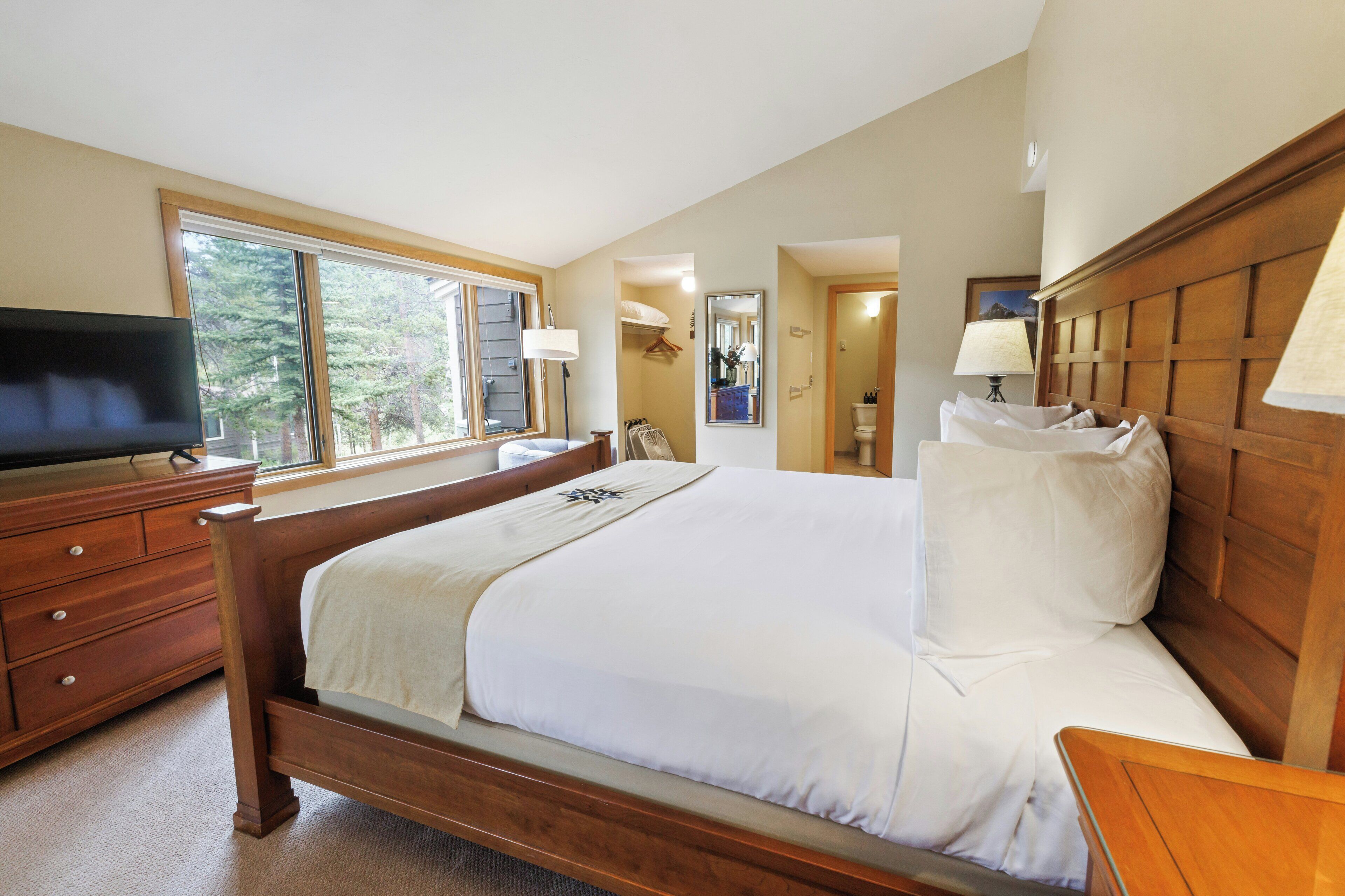A bedroom with a wooden bed, a television on a dresser, and a window with a view of trees.