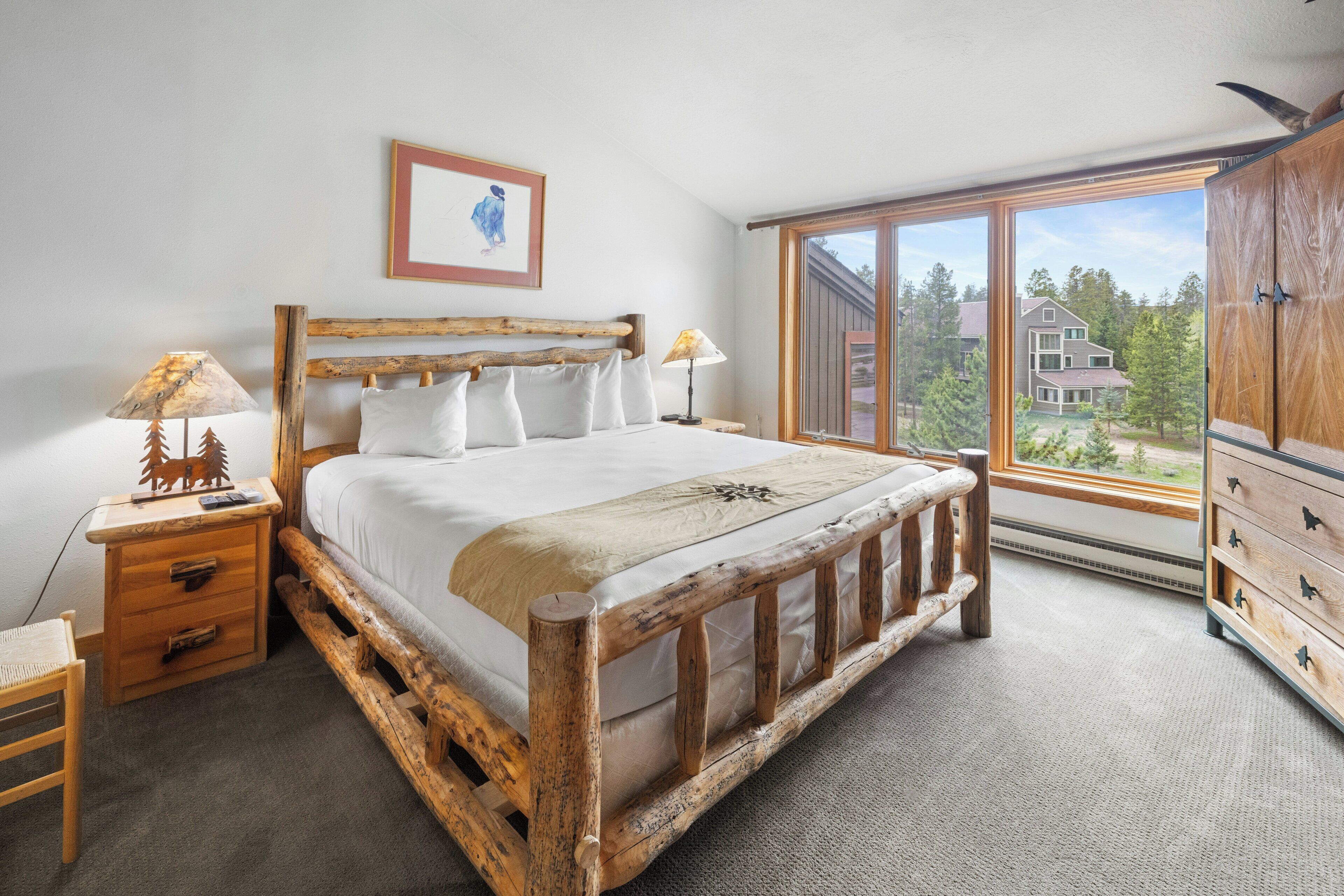 A bedroom with a wooden bed, a nightstand, a dresser, and a large window offering a view of buildings and trees.