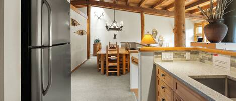 A modern kitchen with wooden cabinets, a stainless steel refrigerator, a dining area with a wooden table and chairs, and a chandelier.