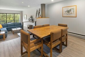 A modern living room with a wooden dining table, cushioned chairs, a blue sofa, and a large window offering a view of trees.