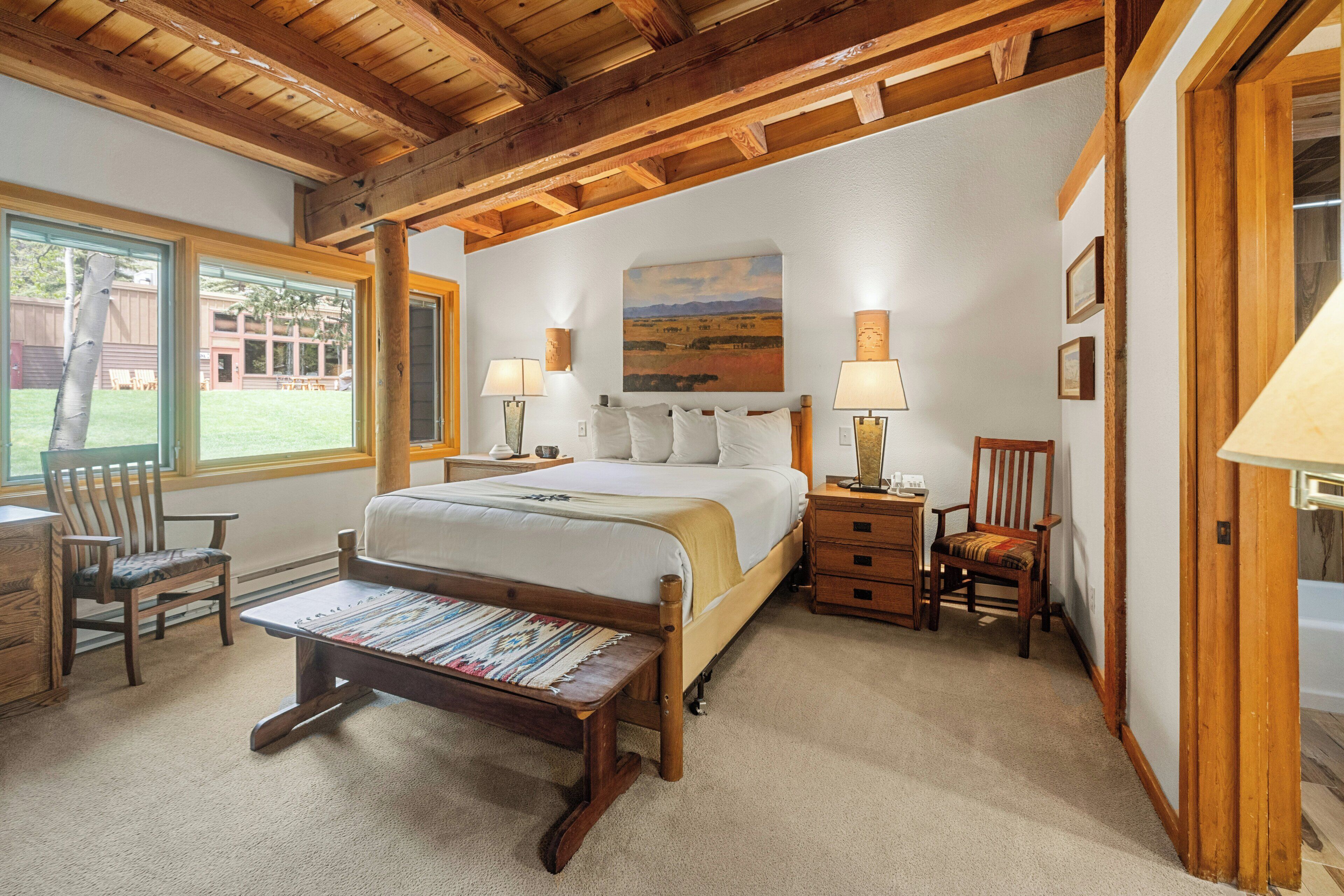A bedroom with a wooden bed, two bedside tables with lamps, a bench, and a view of the outdoors.