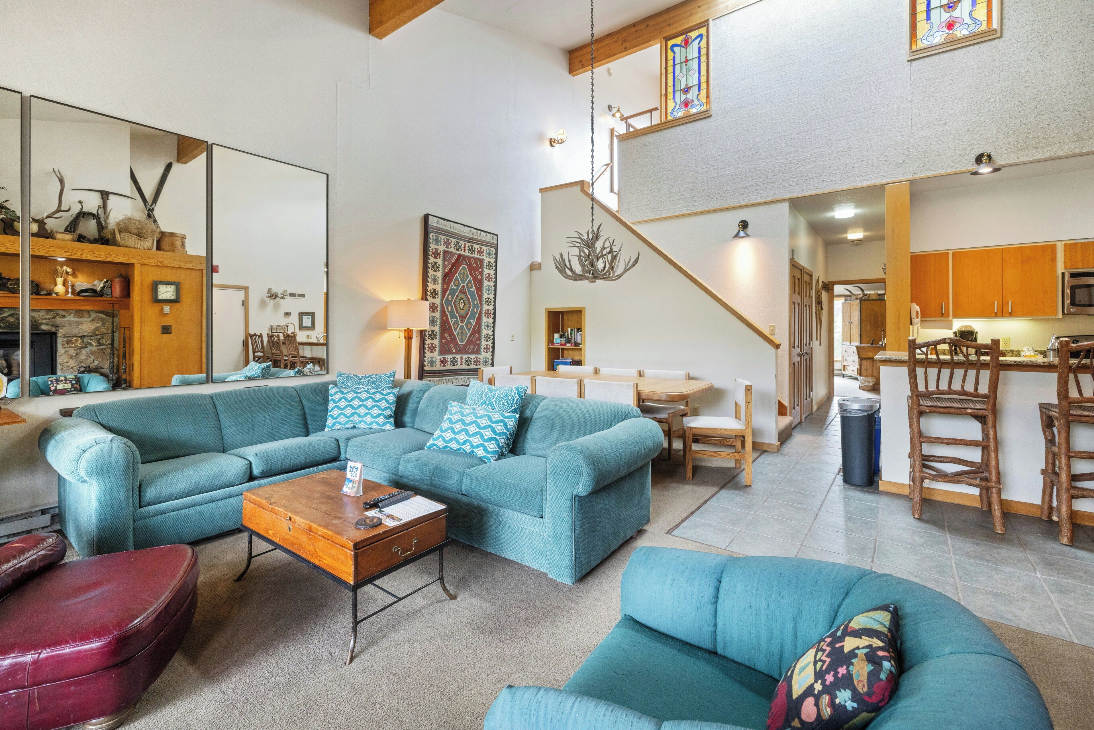A spacious living room with a large blue sofa set, a wooden coffee table, and a red ottoman. A staircase leads to an upper level with a kitchen area visible.