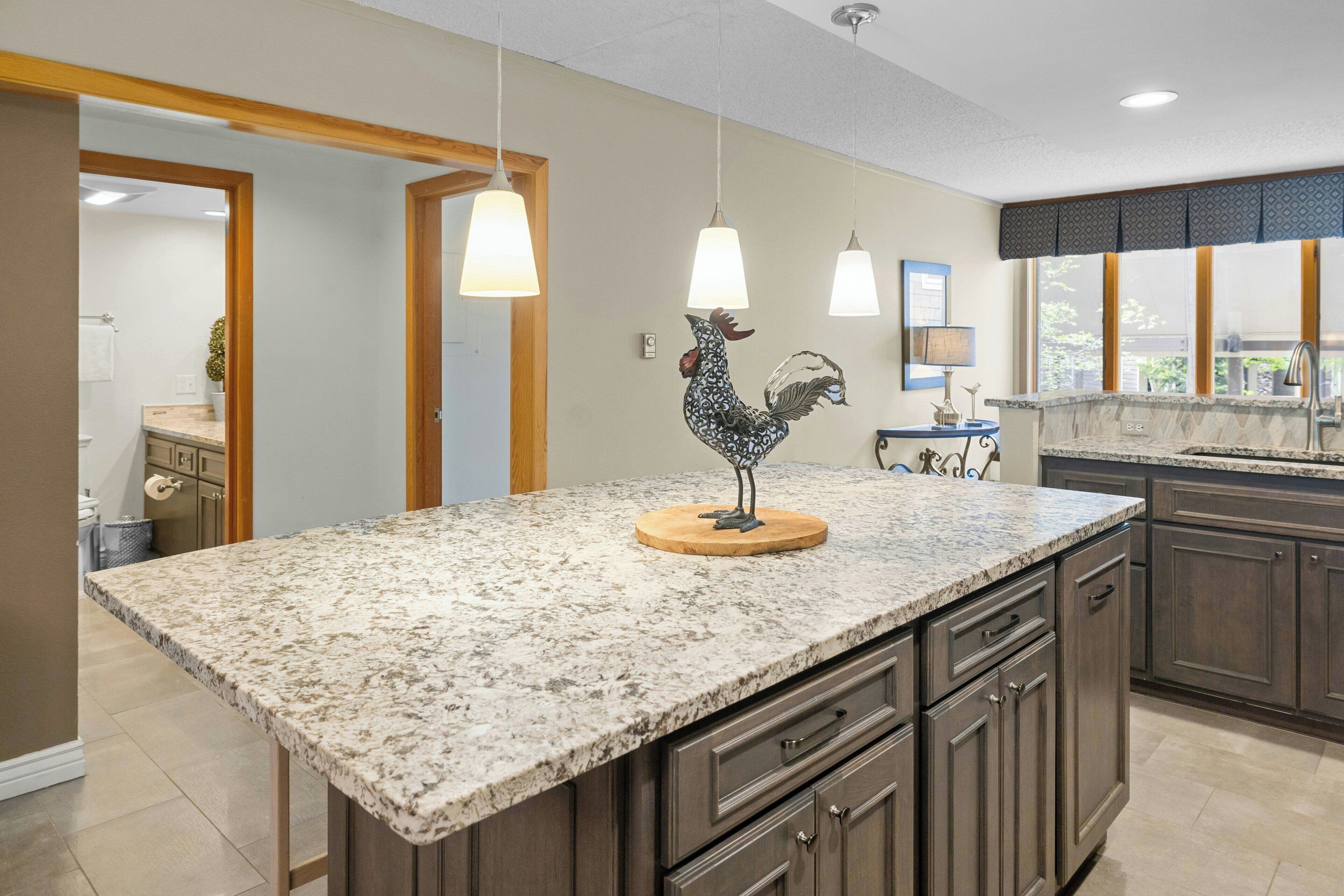 A kitchen with granite countertops, wooden cabinets, pendant lighting, and a rooster decoration.