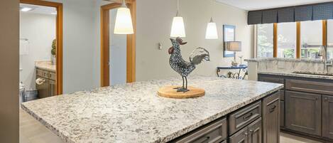 A kitchen with granite countertops, wooden cabinets, pendant lighting, and a rooster decoration.