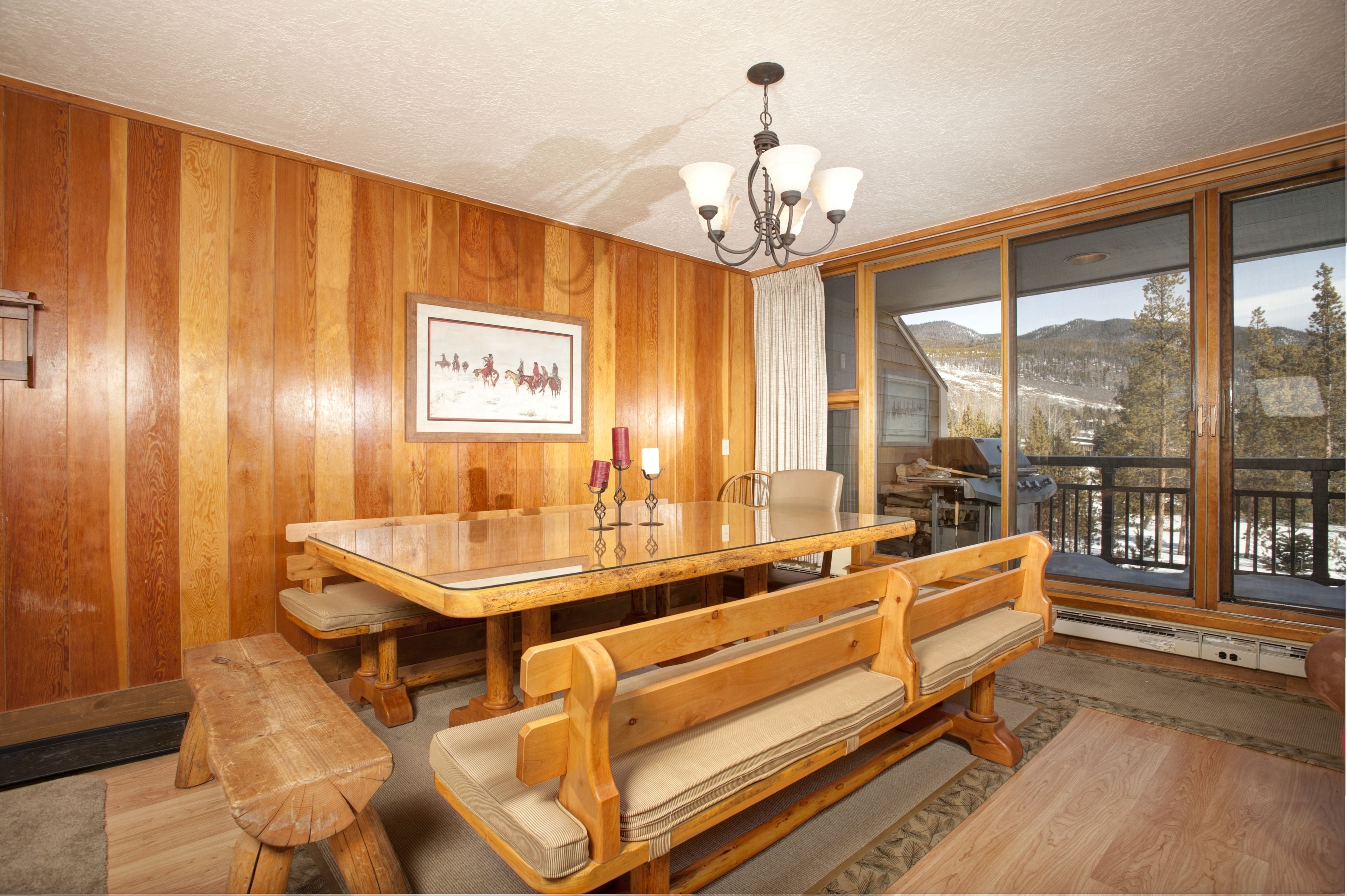 A wooden-paneled room with a large dining table, benches, a chandelier, a framed picture, and a large window with a mountain view.