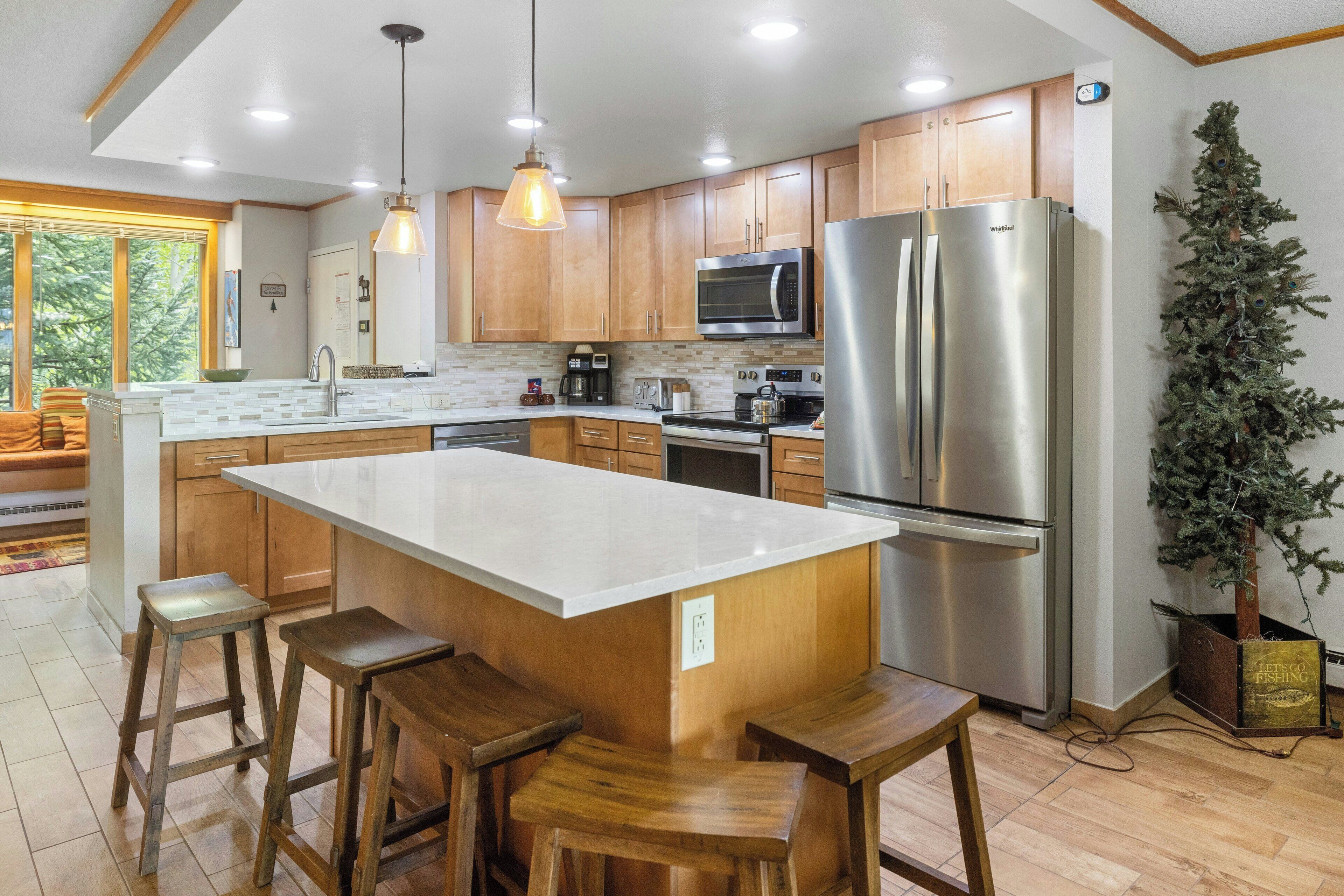 A modern kitchen with wooden cabinets, a large island with stools, stainless steel appliances, and a potted plant.