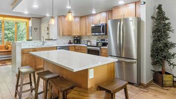 A modern kitchen with wooden cabinets, a large island with stools, stainless steel appliances, and a potted plant.