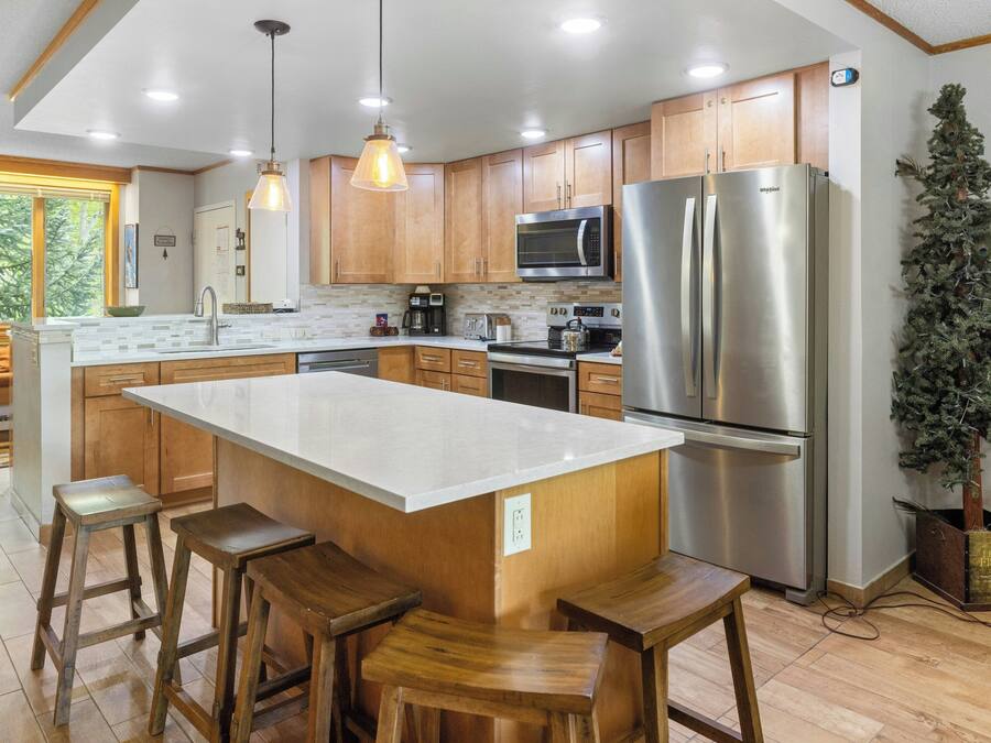 A modern kitchen with wooden cabinets, a large island with stools, stainless steel appliances, and a potted plant.