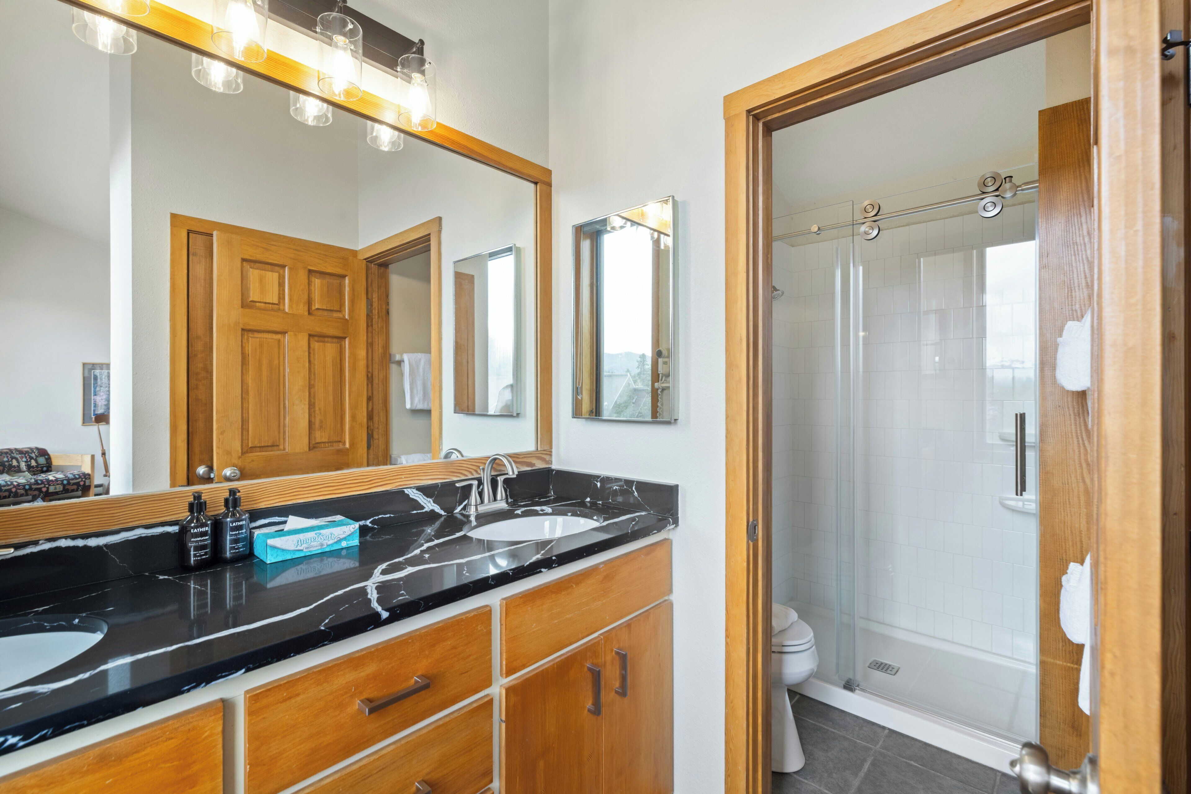 A modern bathroom with a black marble countertop, wooden cabinets, a large mirror, and a glass-enclosed shower.