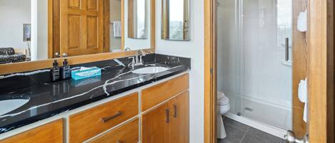 A modern bathroom with a black marble countertop, wooden cabinets, a large mirror, and a glass-enclosed shower.
