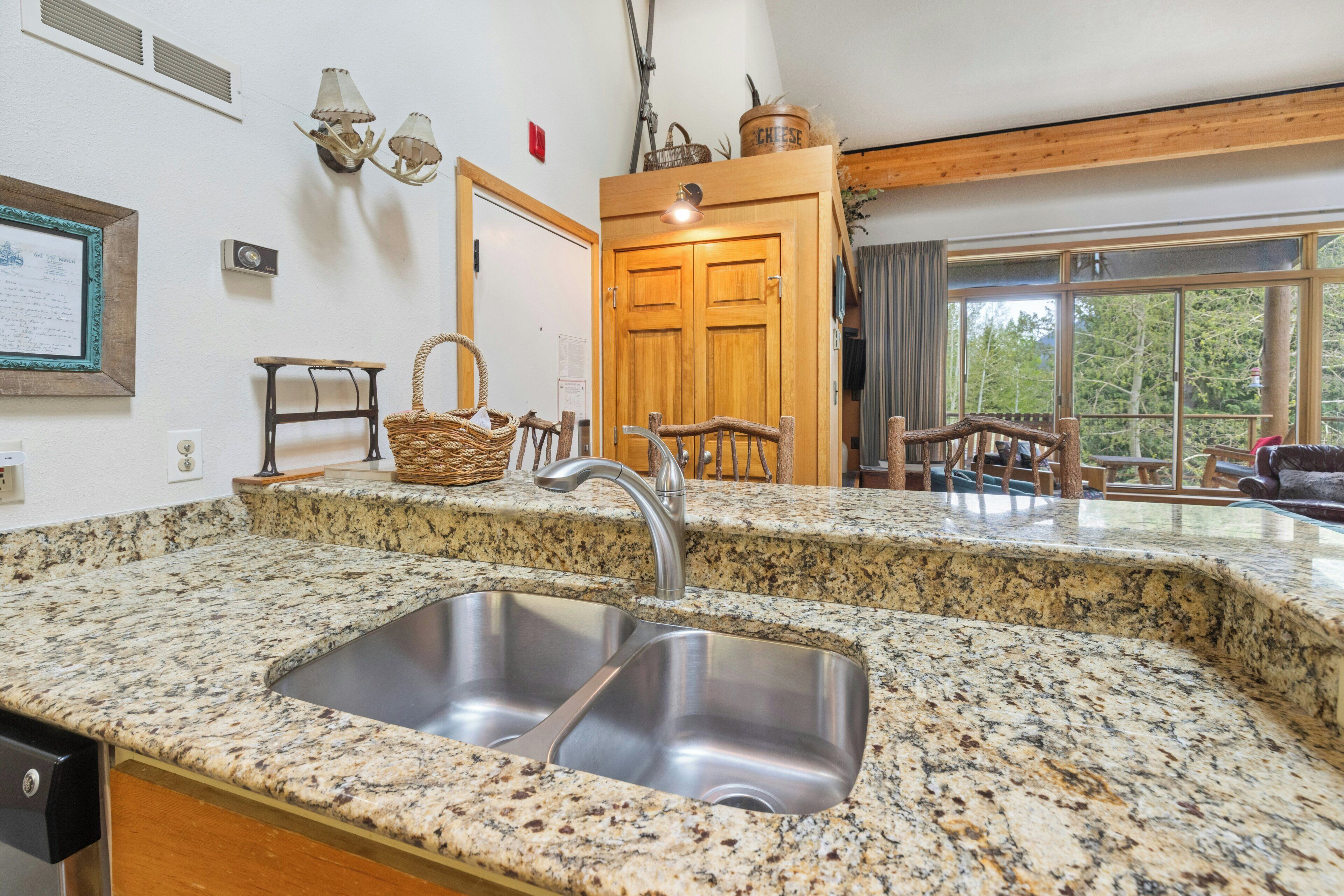 A kitchen with granite countertops, a double stainless steel sink, wooden cabinets, and a wooden door leading to a balcony.