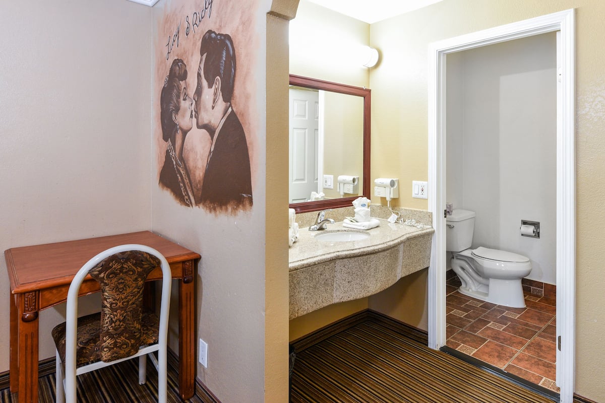 Un baño con lavamanos, espejo e inodoro. Hay una silla y una mesa con un adorno encima.