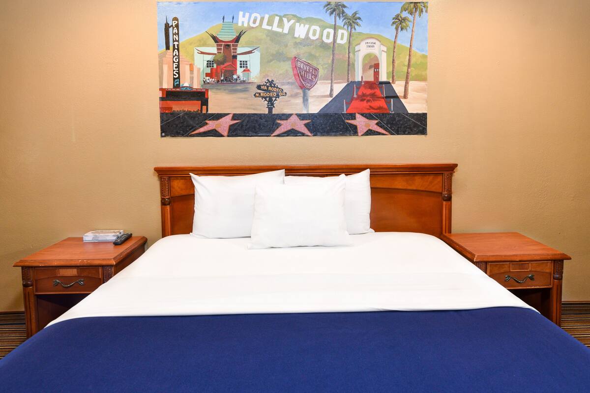 Habitación de hotel con cama, dos mesitas de noche, un control remoto y un cuadro enmarcado de Hollywood en la pared.