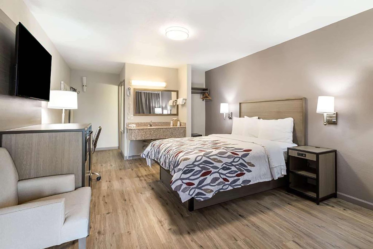 A hotel room with a bed, bedside table, chair, and a desk with a flat-screen TV.