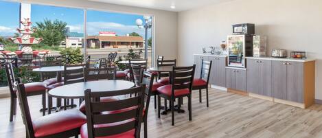 A restaurant with round tables and chairs, a kitchen area, and large windows offering a view of the outside.