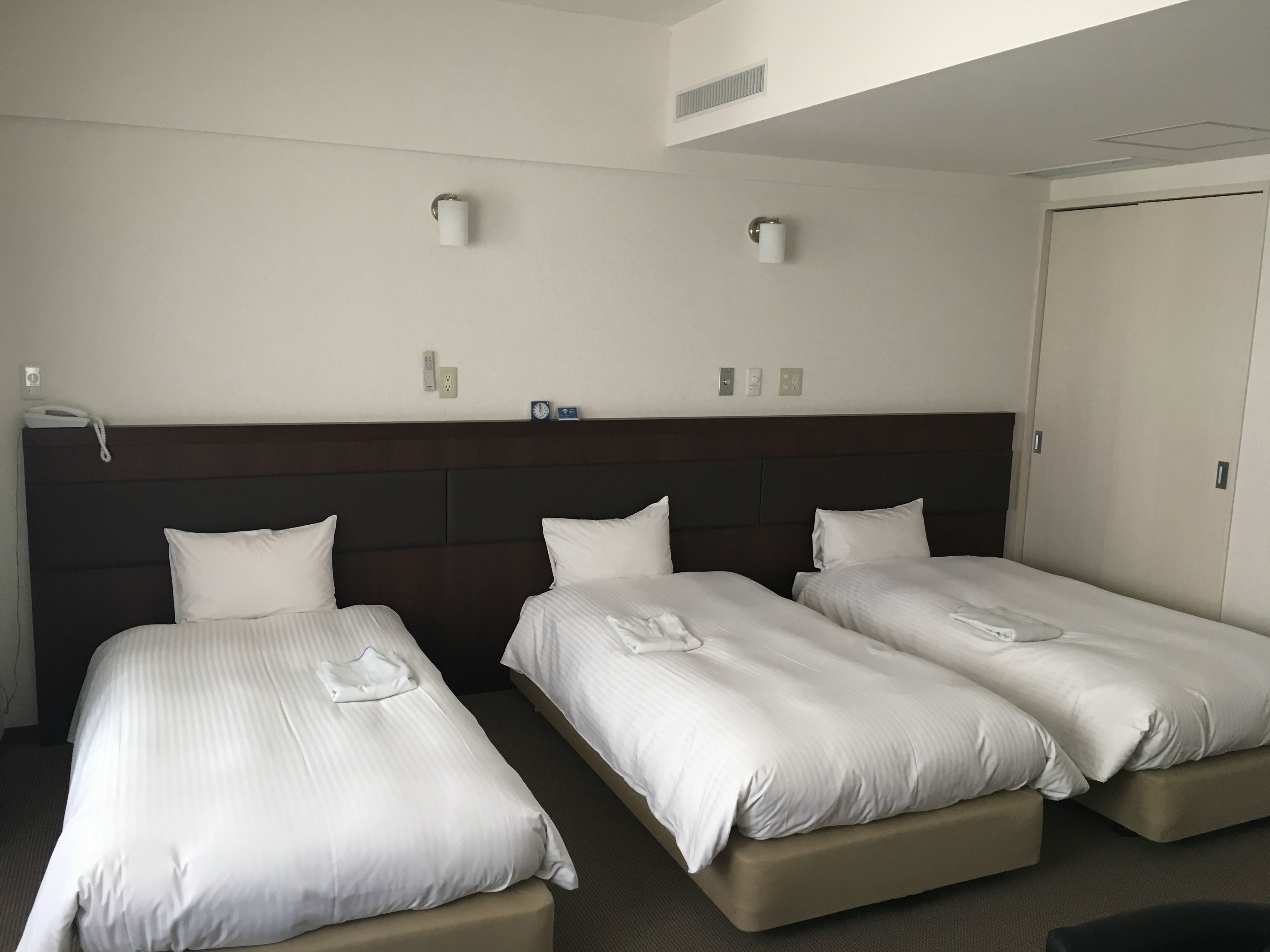 Une chambre d’hôtel avec trois lits simples, une tête de lit sombre, un mur blanc et une porte.
