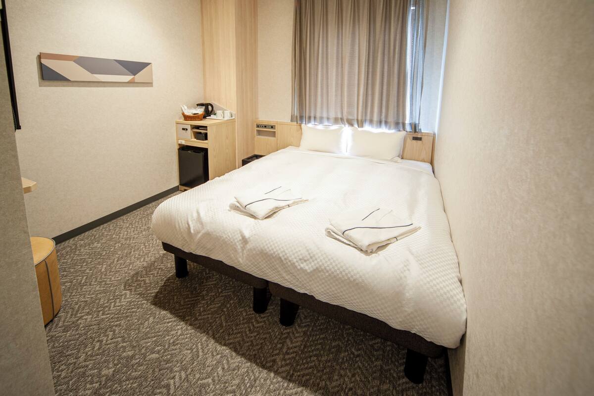 Een hotelkamer met een groot bed, een bureau met koffiezetapparaat, een klein kastje en een raam met gordijnen.
