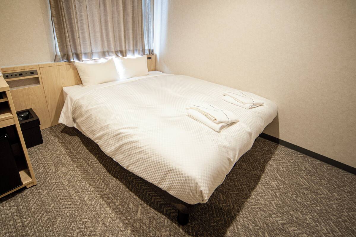 Een net opgemaakt bed met twee opgevouwen handdoeken, een houten nachtkastje en een prullenbak.