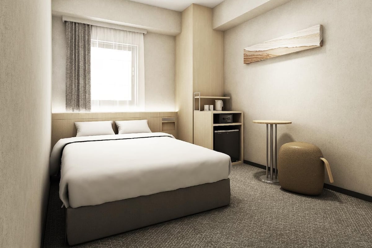 Quarto de hotel com cama, mesa pequena, cadeira, guarda-roupa e janela com cortinas.