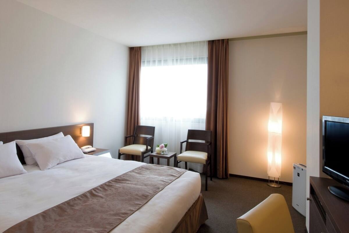 A hotel room with a large bed, two chairs, a small table with a vase, a TV, and a window with curtains.