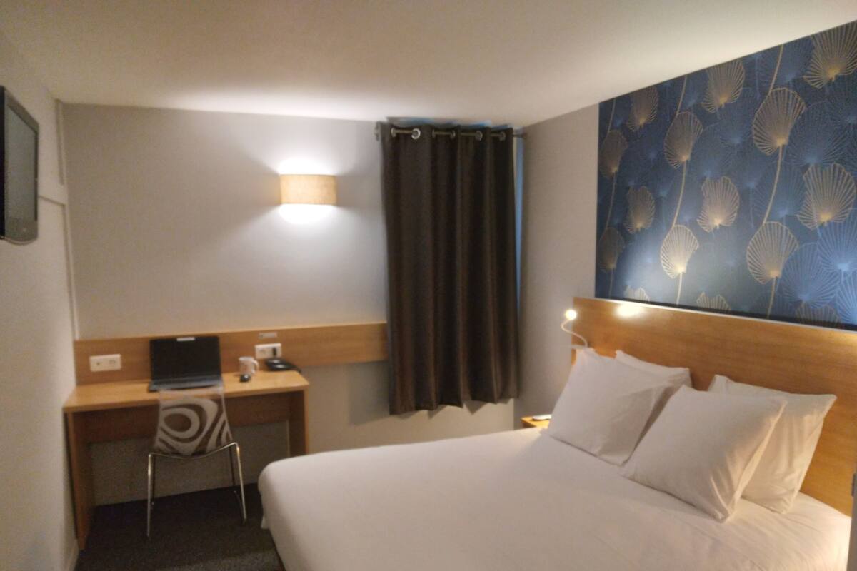 Une chambre d’hôtel comprenant un lit, un bureau, une chaise, une lampe et un mur orné d’un motif floral.