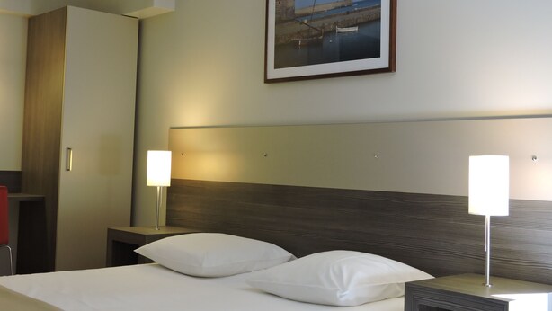 Ein Hotelzimmer mit einem Bett, zwei Nachttischlampen, einem Kopfteil, einem gerahmten Bild an der Wand und einem Kleiderschrank.