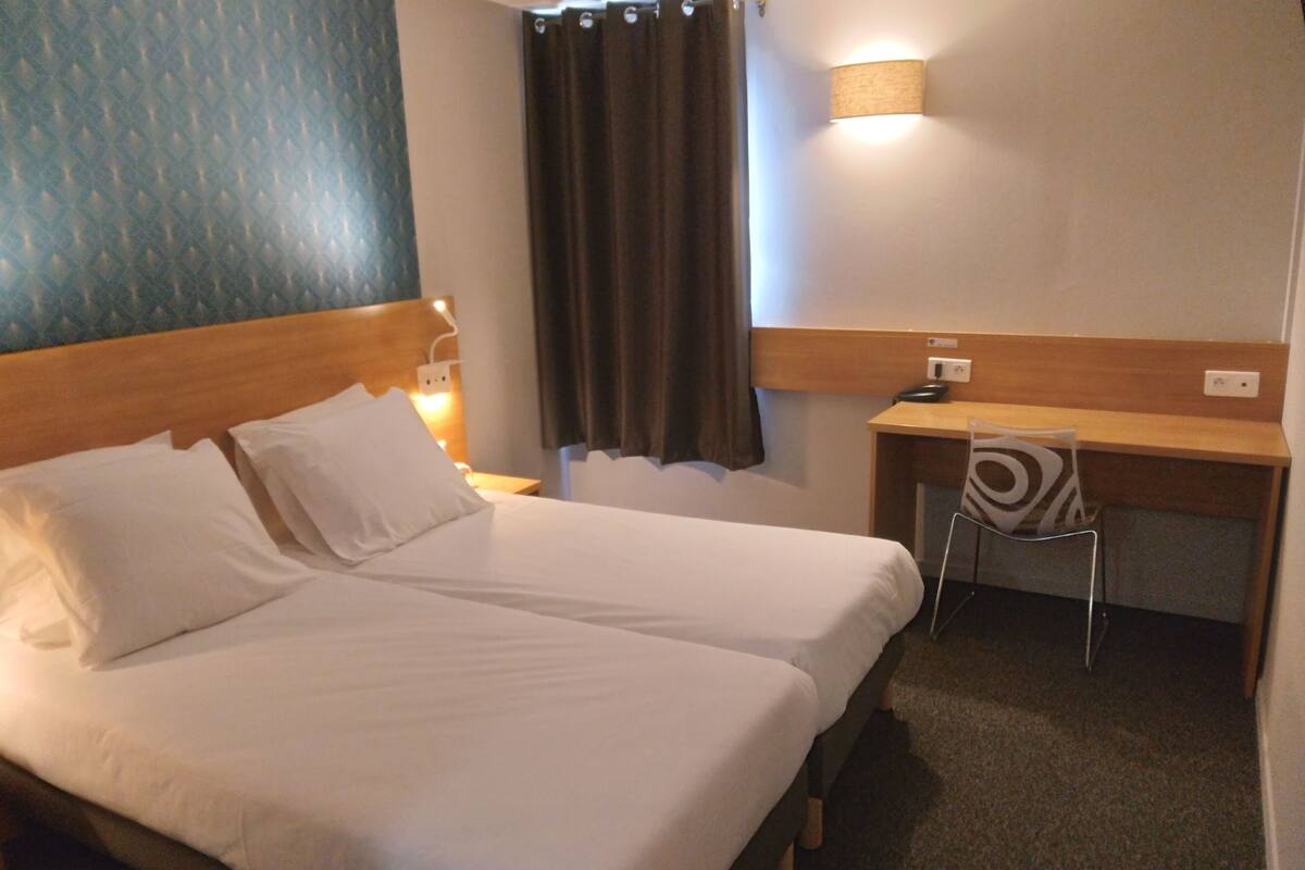 Une chambre d’hôtel avec deux lits, un bureau, une chaise, une lampe et un rideau.
