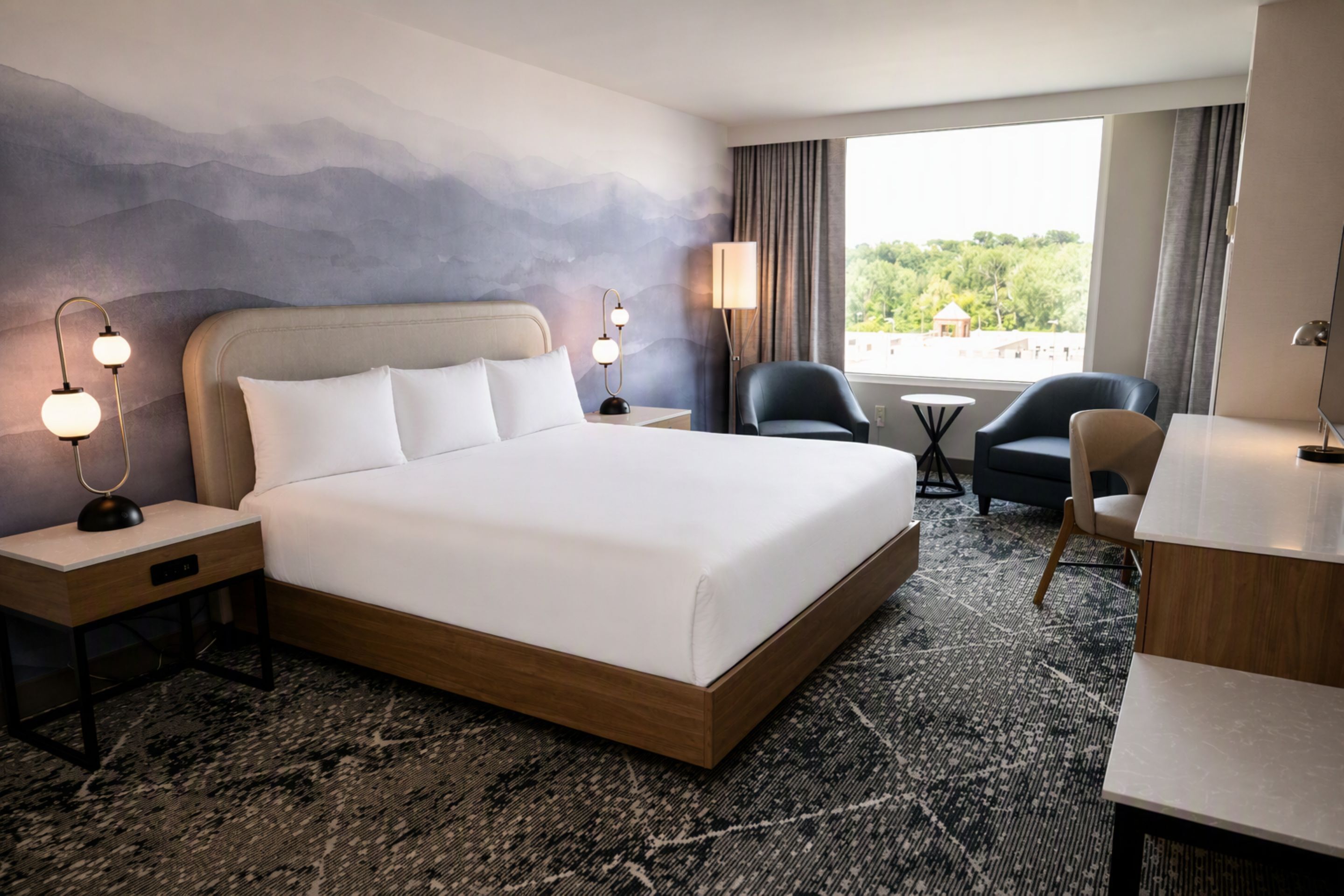A hotel room with a large bed, two bedside tables, a desk, a chair, and a view of the outdoors.
