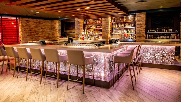 A bar area with a long counter, multiple bar stools, and shelves stocked with bottles. - Harrah's Kansas City Hotel & Casino - A Caesars Rewards Destination (North Kansas City)