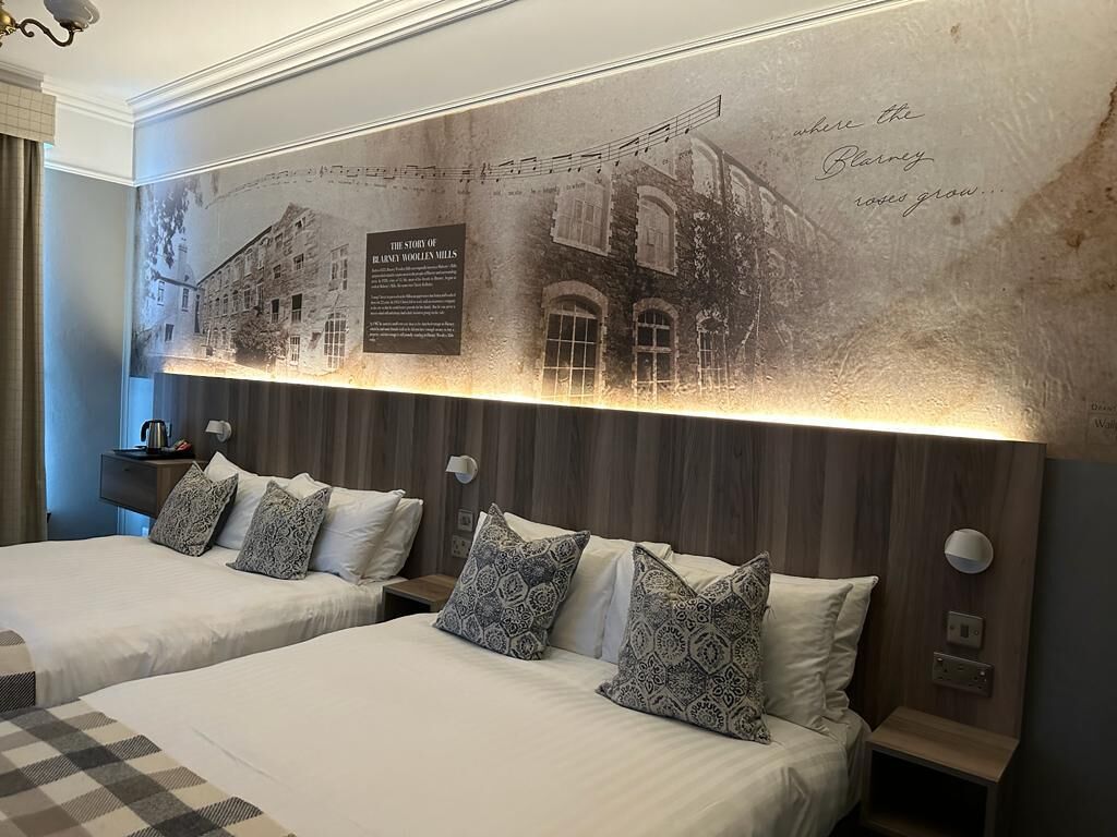 Habitación de hotel con dos camas, almohadas estampadas, una lámpara de pared y un mural histórico.
