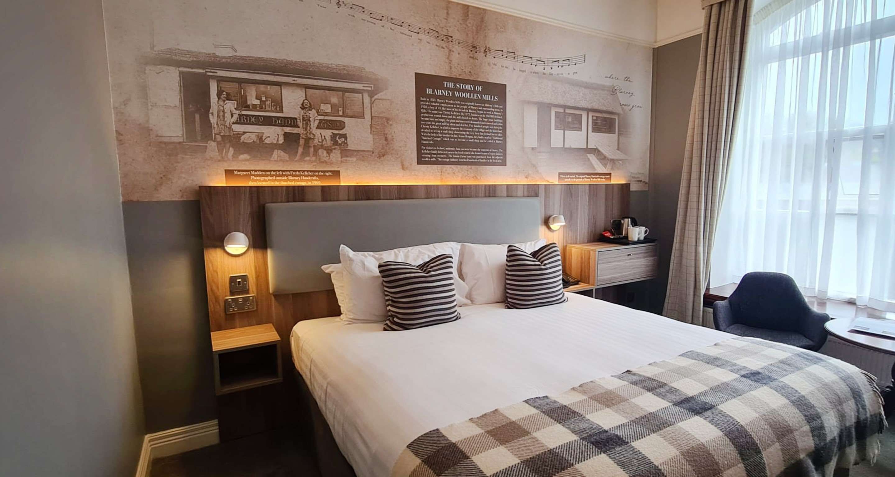 A hotel room with a large bed, two bedside tables, a nightstand, a desk, and a chair. There is a window with curtains, a wall-mounted clock, and a plaque above the bed.