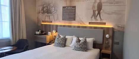 A hotel room with a large bed, two pillows, a nightstand, a desk, and a wall with a poster.