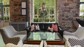 A room with a stone wall, a large arched window, a black leather sofa, patterned armchairs, and a glass coffee table.