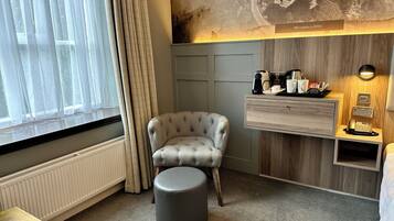 Habitación de hotel con un sillón capitonado, un pequeño reposapiés, una mesita de noche de madera con un teléfono y una lámpara, y una estantería con una cafetera y tazas.