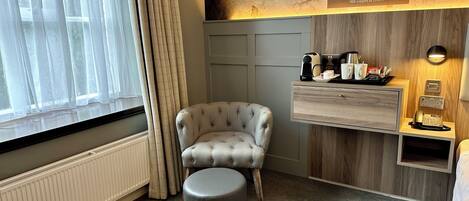 Habitación de hotel con un sillón capitonado, un pequeño reposapiés, una mesita de noche de madera con un teléfono y una lámpara, y una estantería con una cafetera y tazas.