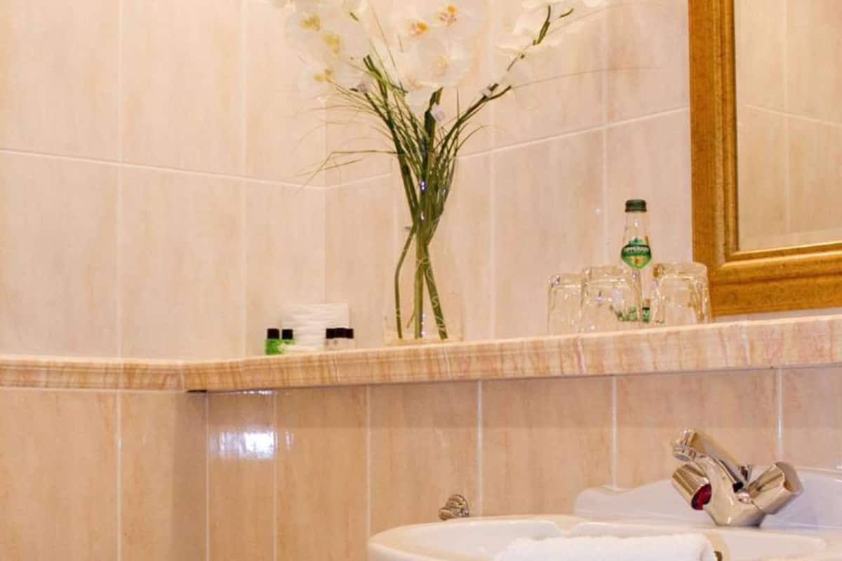 Un baño con pared de mármol, un lavamanos con espejo, un dispensador de papel higiénico y un jarrón con flores.