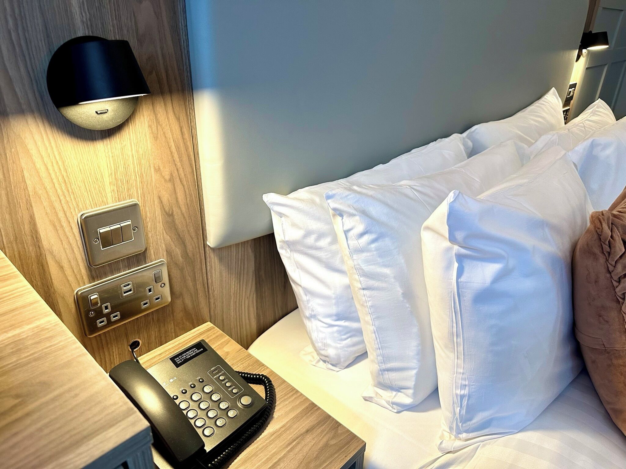 A hotel room with a wooden headboard, a bedside telephone, a wall-mounted light, and a bed with white pillows.