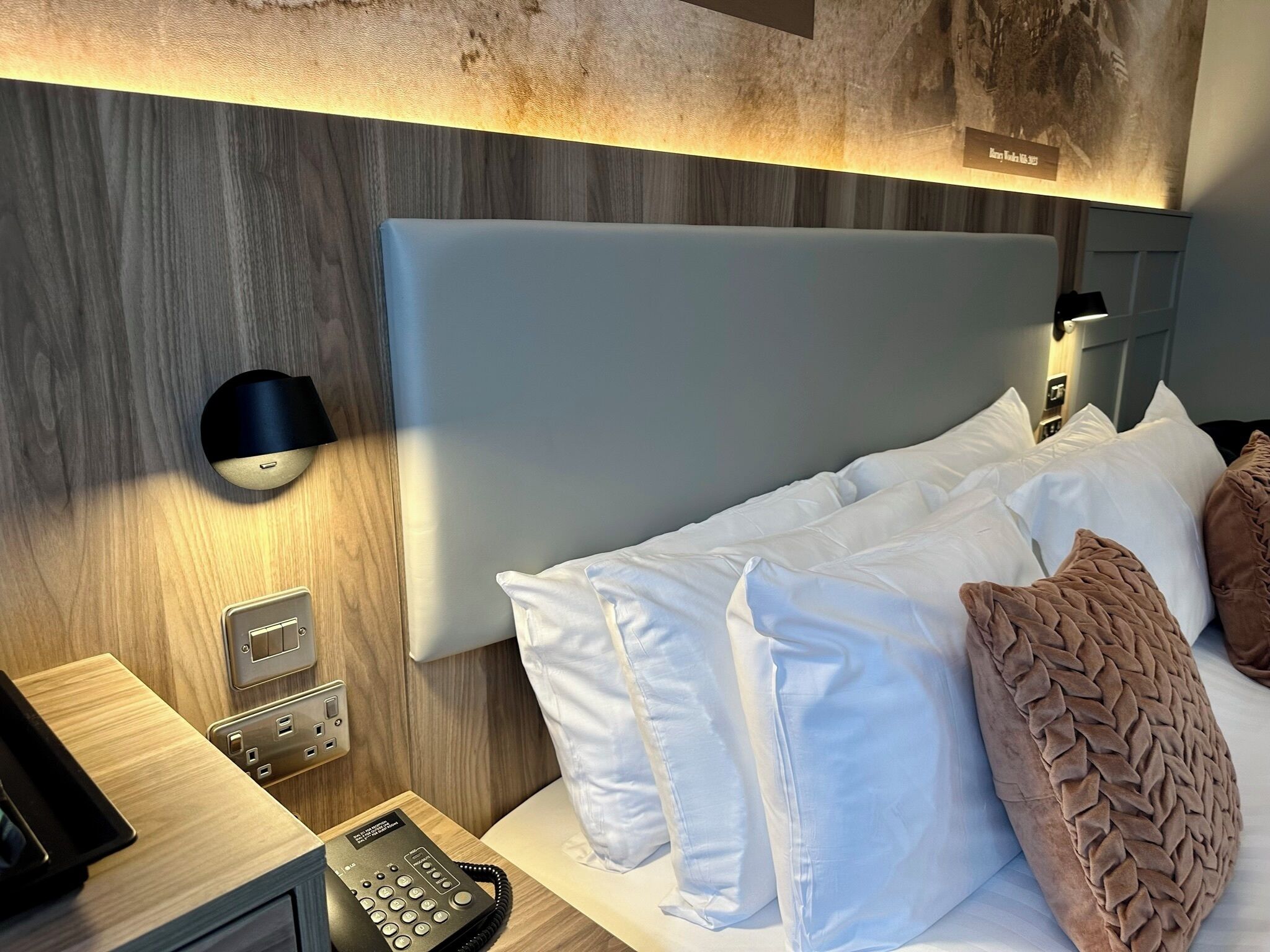 Una habitación de hotel con cabecera de madera, teléfono en la mesita de noche y lámpara de pared.