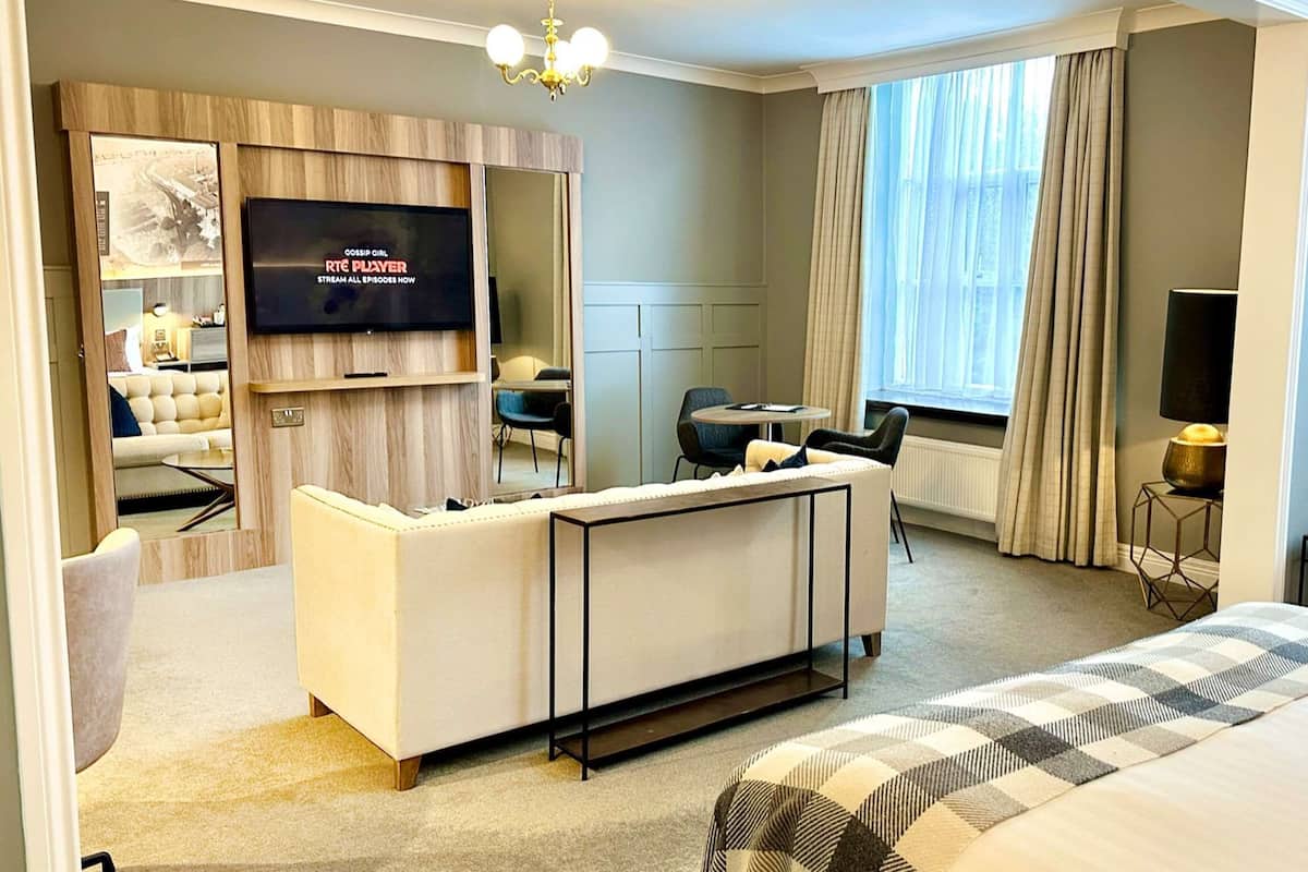 Una habitación de hotel con una cama grande, un televisor montado en la pared, una zona de estar con un sofá y sillones, y una mesita.